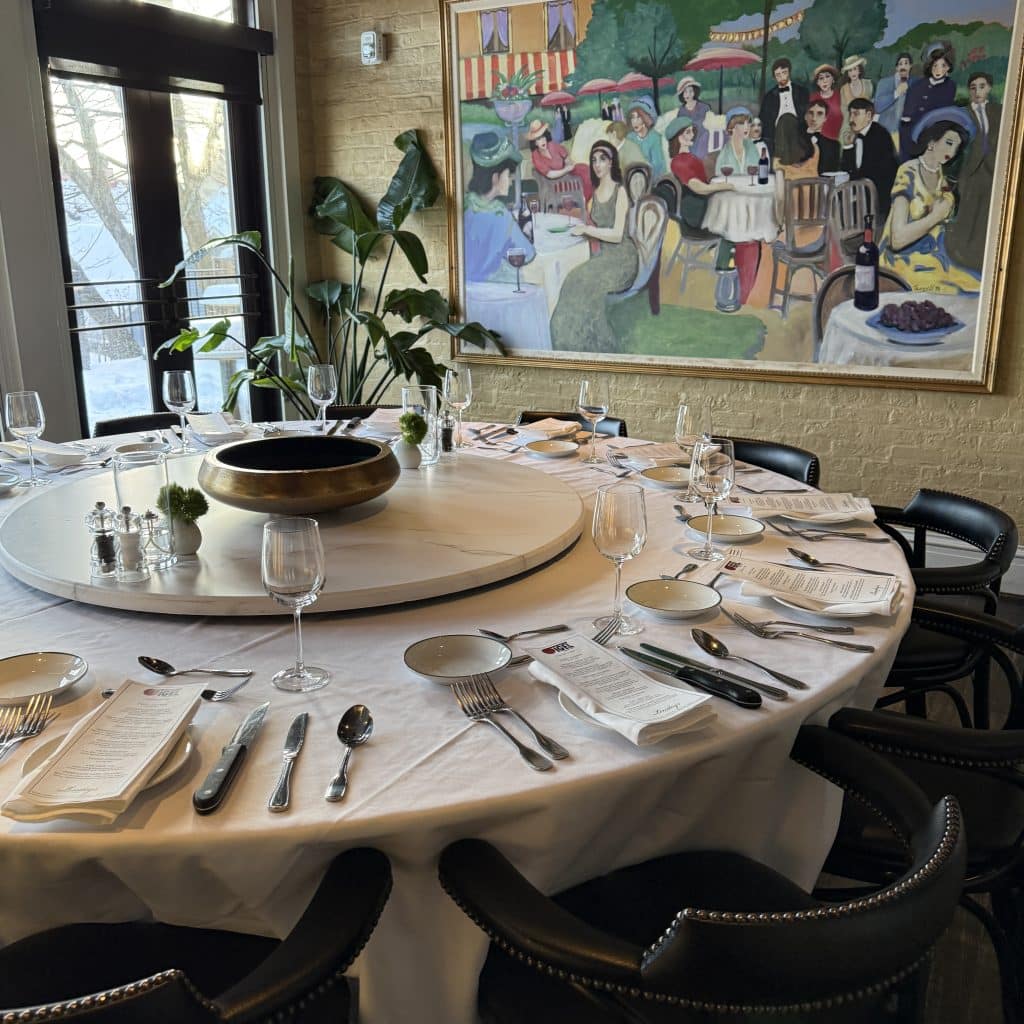 A circular dining table organized with detailed table setting including a drinking glass, napkins, plate, and silverware